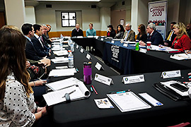 A group of people are sitting a table facing each other. Materials can be seen on the table. 